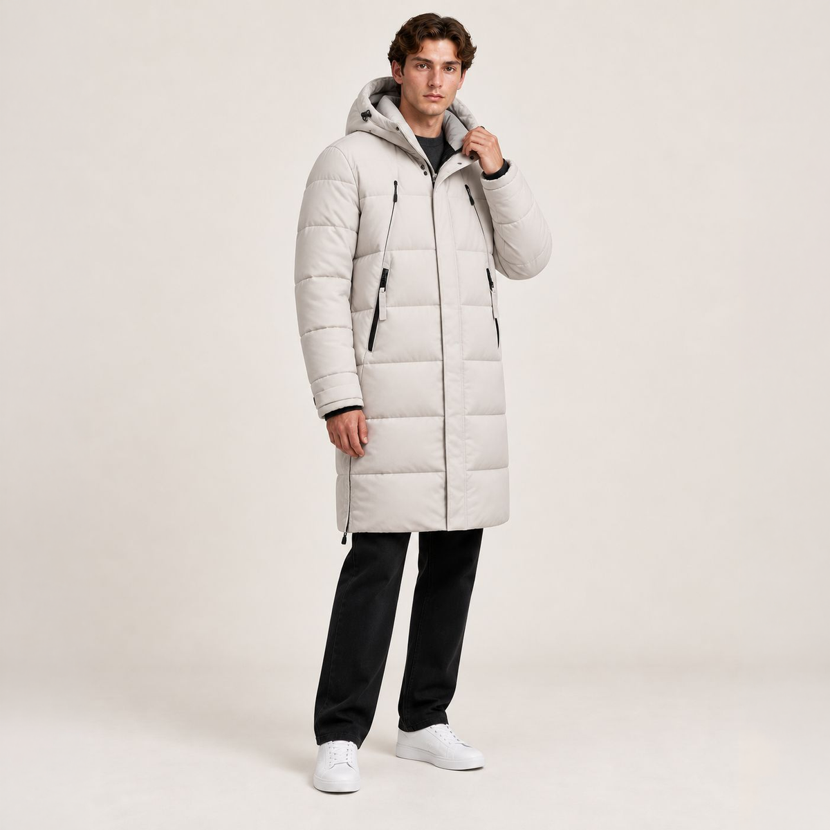 Parka longue beige matelassée à capuche, portée de trois-quarts avec une main au col dans le studio IVAREE, format carré 1:1.