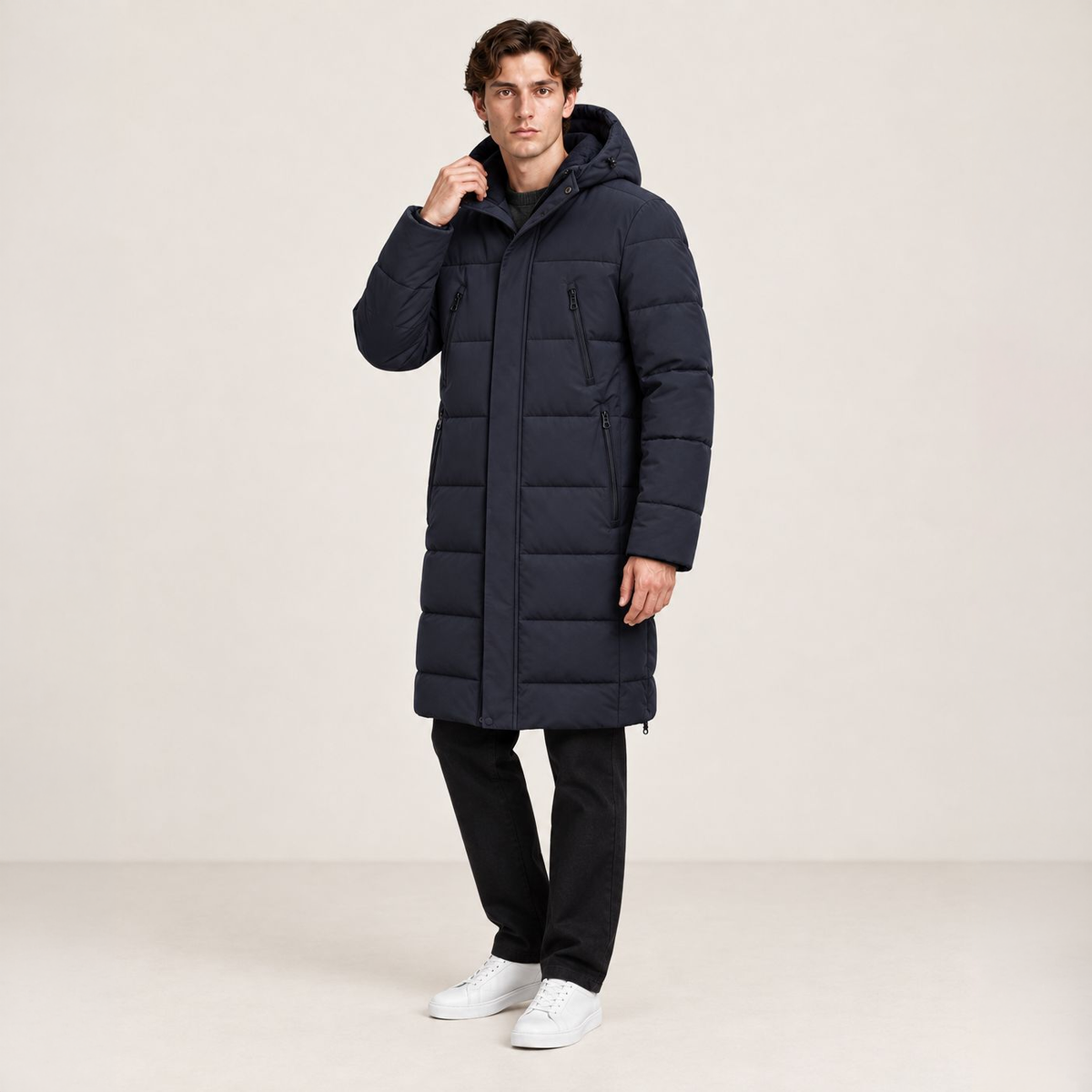 Parka longue bleu foncé matelassée à capuche, portée de trois-quarts avec une main au col dans le studio IVAREE, format carré 1:1.