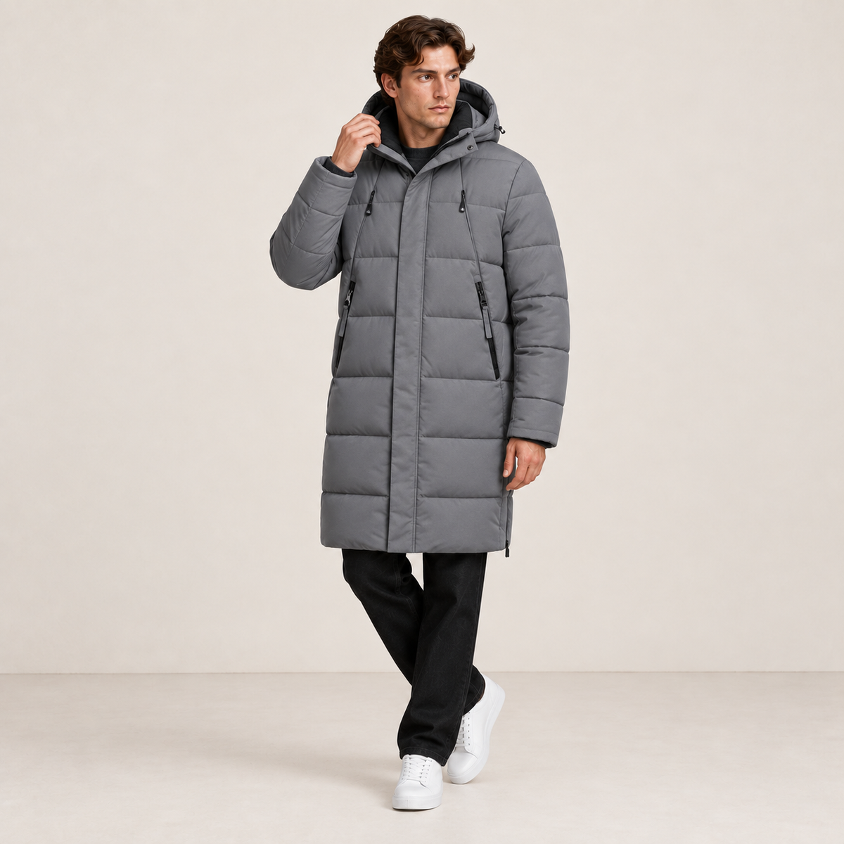 Parka longue grise matelassée à capuche, portée de trois-quarts avec une main au col dans le studio IVAREE, format carré 1:1.