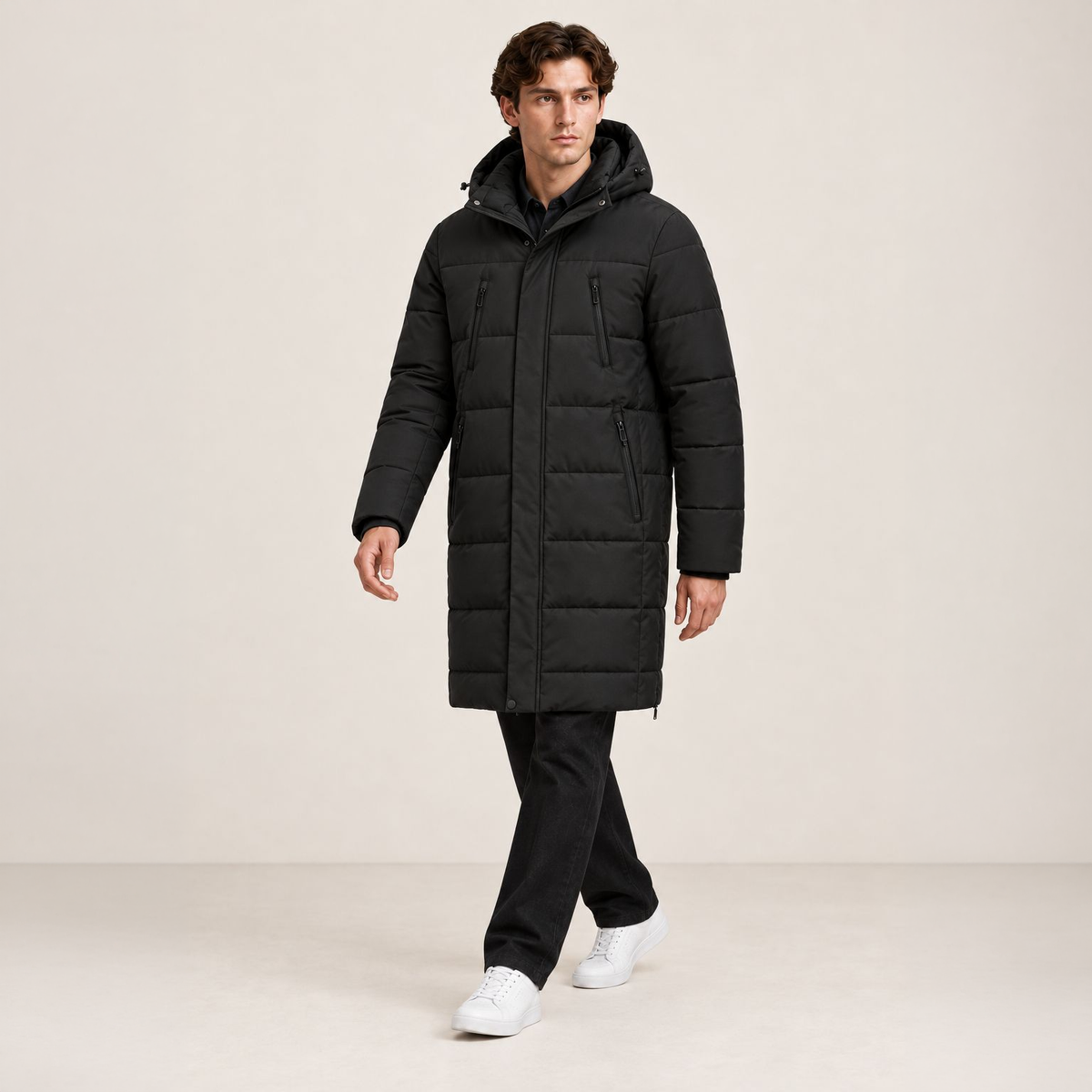 Parka longue noire matelassée à capuche et quatre poches zippées, portée en marche dans le studio IVAREE, format carré 1:1.
