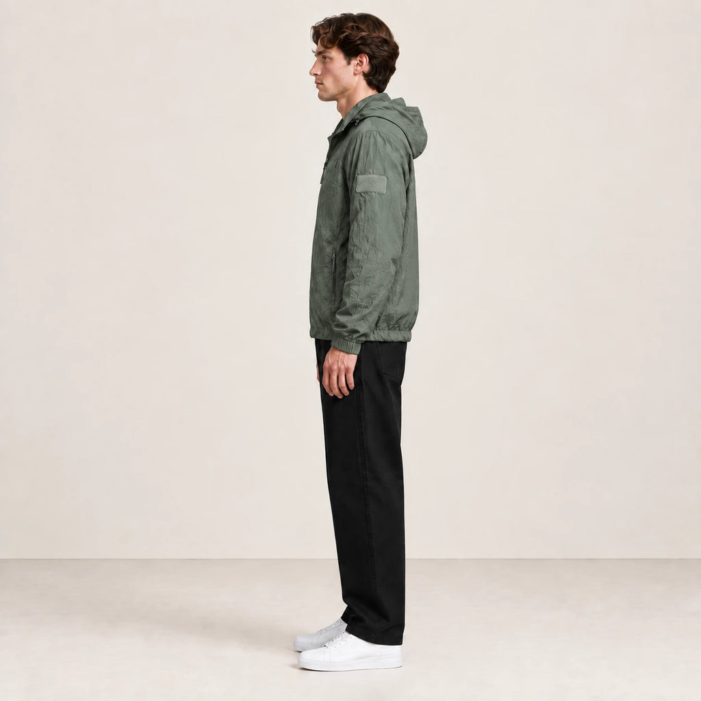 Veste légère vert grisé à capuche et fermeture zippée, portée de profil dans le studio IVAREE.