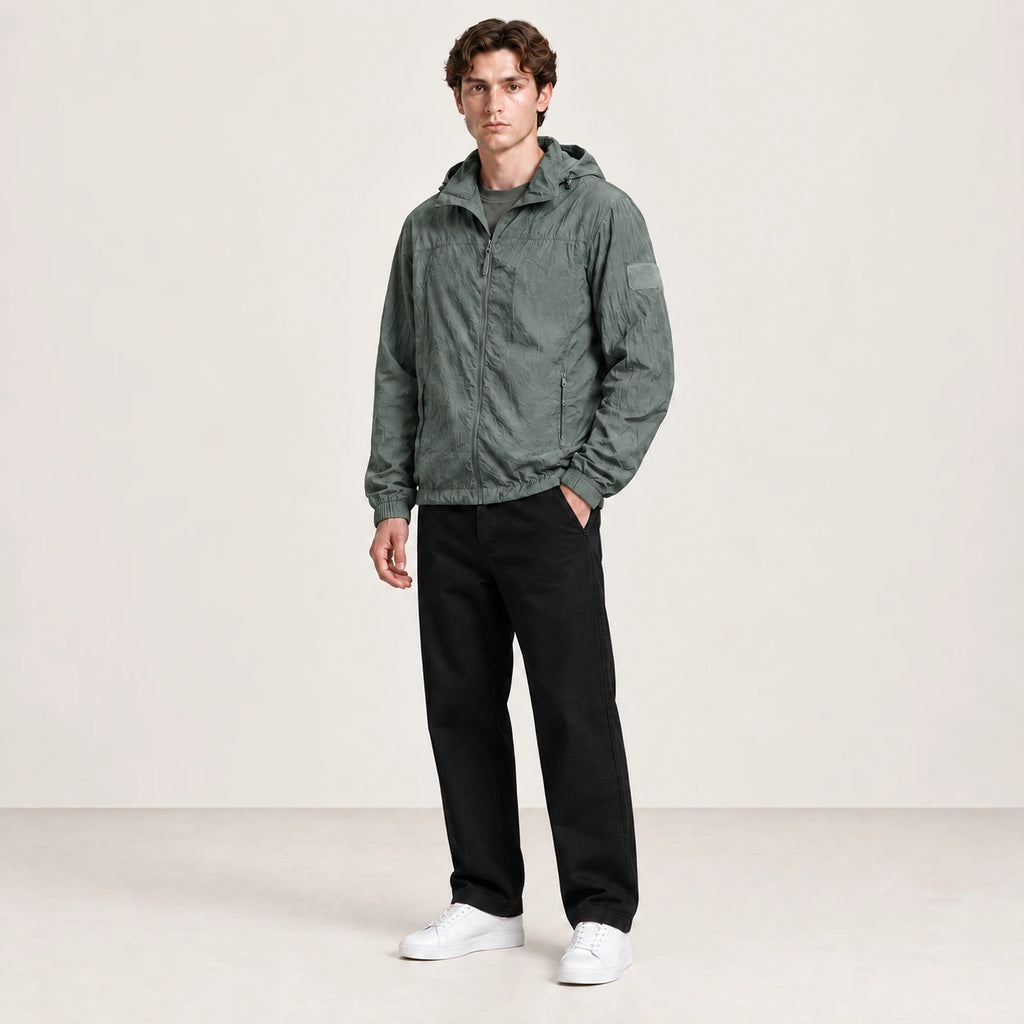 Veste légère vert grisé à capuche et fermeture zippée, portée de trois-quarts dans le studio IVAREE.